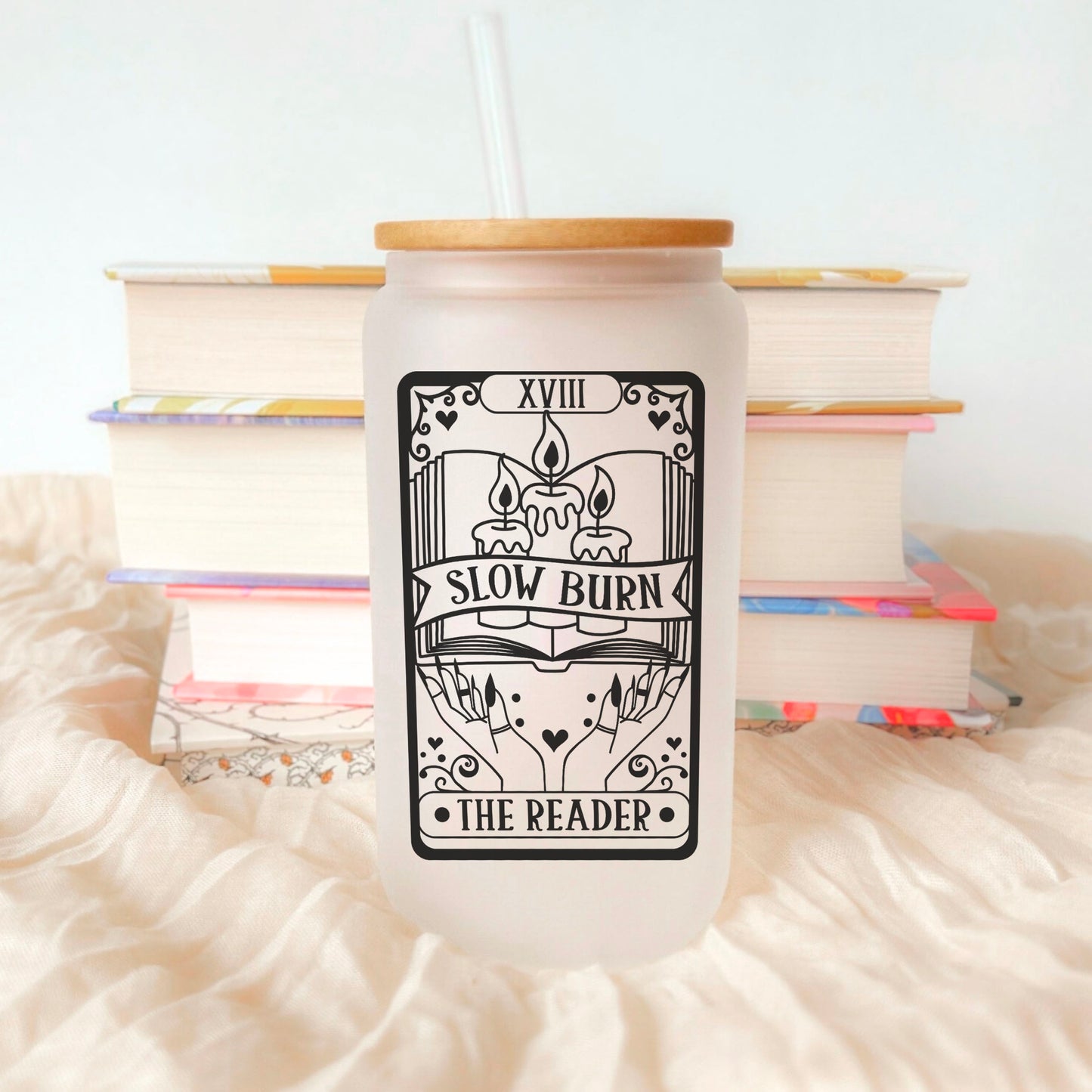 A frosted glass cup with a bamboo lid and glass straw featuring a tarot-style bookish design with melting candles, an open book, and the words "Slow Burn - The Reader." Set against a cozy backdrop with stacked books, this aesthetic drinkware is perfect for book lovers who adore slow-burn romance and literary-inspired accessories. Ideal for iced coffee, tea, or any favorite beverage, this glass cup makes a charming addition to any reader’s collection.